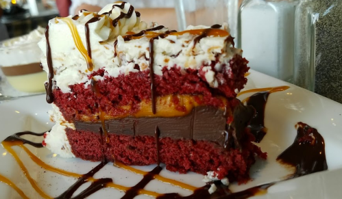 A slice of red velvet cake layered with chocolate and caramel, topped with whipped cream and drizzled with chocolate sauce.