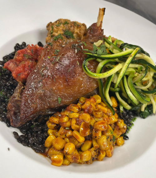 A plated dish featuring duck leg, lentils, corn, tomato sauce, and zucchini noodles, garnished with herbs.