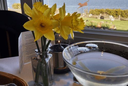 A vase of yellow daffodils on a table with a candle and a martini, overlooking a scenic waterfront view.