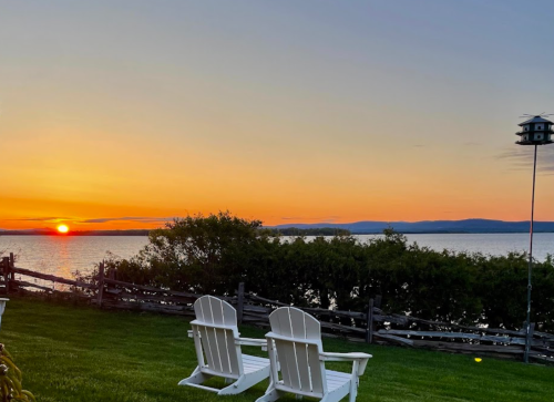 Two white Adirondack chairs overlook a tranquil lake at sunset, with a colorful sky and distant mountains in the background.
