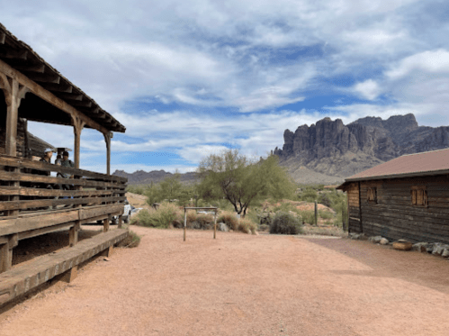 A rustic wooden structure with a porch, surrounded by desert landscape and mountains under a cloudy sky.