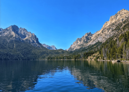 A serene lake surrounded by mountains and lush forests under a clear blue sky. Reflections shimmer on the water's surface.