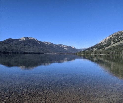 A serene lake surrounded by mountains under a clear blue sky, with calm water reflecting the landscape.