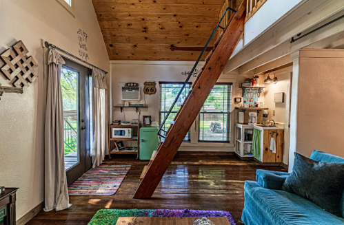 Cozy interior of a small cabin with wooden beams, a blue couch, and a kitchenette, featuring large windows and natural light.