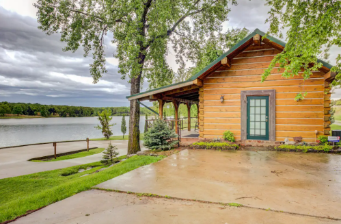 A rustic log cabin by a lake, surrounded by greenery and trees, under a cloudy sky.