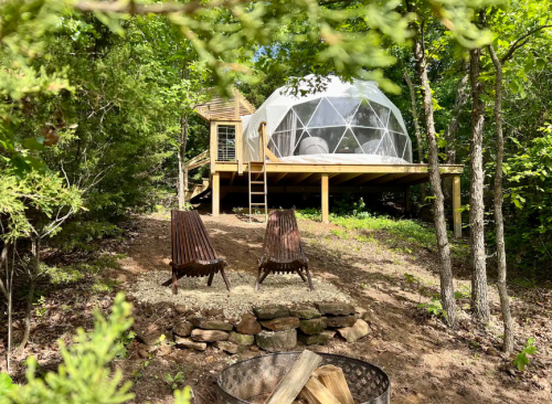 A geodesic dome cabin on a wooden deck surrounded by trees, with two chairs and a fire pit in the foreground.
