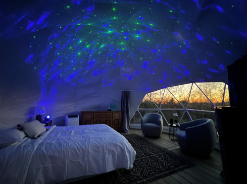 A cozy dome interior with a bed, two chairs, and a starry light projection on the ceiling at sunset.