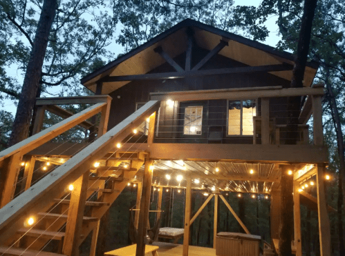 A cozy treehouse with warm lights, wooden stairs, and a balcony surrounded by trees at dusk.