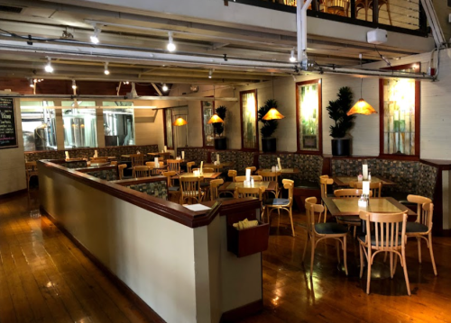 A cozy restaurant interior with wooden floors, tables, and soft lighting, featuring plants and a bar area in the background.