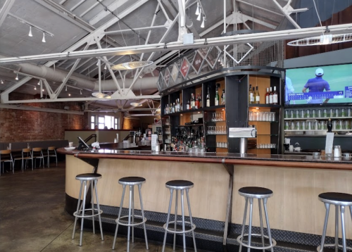 A modern bar with a wooden counter, high stools, and a large TV, set in a spacious, industrial-style interior.