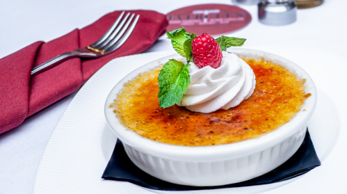 A creamy crème brûlée topped with whipped cream, a raspberry, and mint, served on a white plate with a fork.