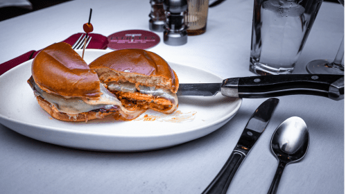 A close-up of a sliced sandwich with melted cheese, served on a white plate with utensils and a glass of water in the background.