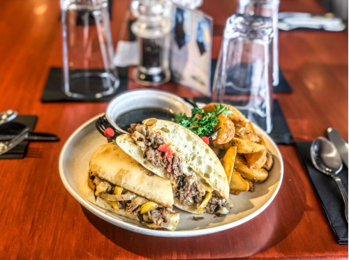 A plate featuring a sandwich filled with meat and vegetables, accompanied by seasoned potato wedges and a dipping sauce.