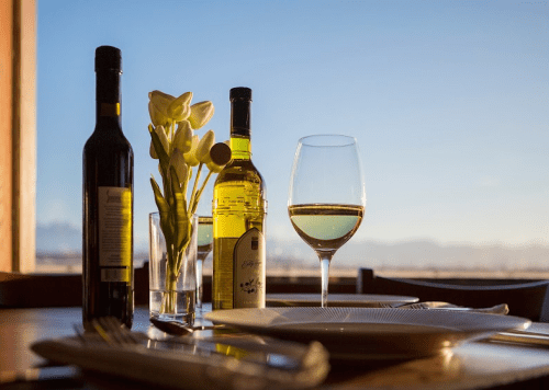 A table setting with a glass of white wine, two bottles, and a vase of flowers against a scenic backdrop.