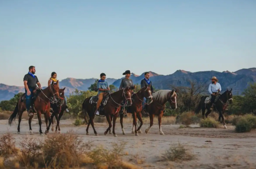 A group of six people on horseback riding through a desert landscape with mountains in the background.