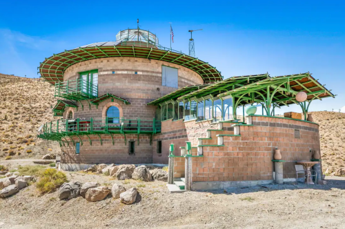 A unique round building with green accents, surrounded by rocky terrain and clear blue skies.
