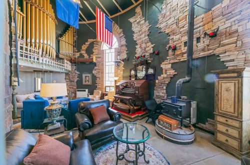 Cozy living room with stone walls, vintage decor, an organ, and flags, featuring a glass coffee table and comfortable seating.