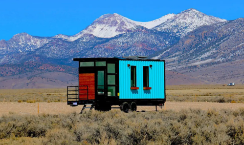 A colorful tiny house on wheels in a vast desert landscape, with mountains in the background under a clear blue sky.
