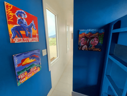 A hallway with blue walls featuring colorful paintings on the left and right, with windows letting in natural light.