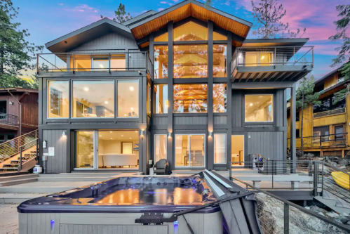 Modern lakeside house with large windows, a hot tub, and a scenic backdrop of trees and sunset.