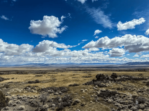 A vast landscape with rolling hills, sparse vegetation, and a blue sky filled with fluffy white clouds.