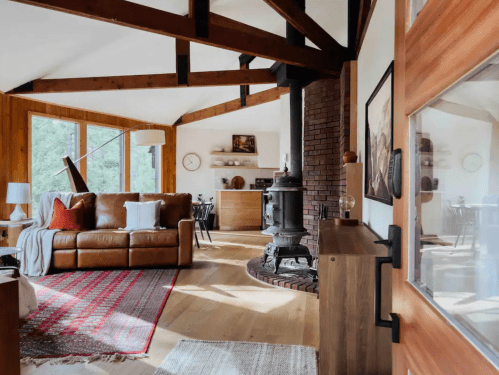 Cozy living room with a brown leather sofa, wooden beams, a vintage stove, and large windows letting in natural light.