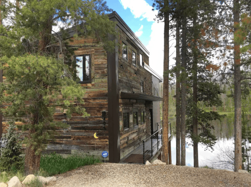 A modern wooden house surrounded by trees, near a calm body of water under a blue sky.