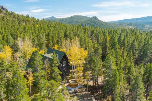 Aerial view of a cabin surrounded by lush green trees and mountains in the background, with a hot tub in the foreground.