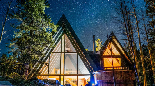 A modern A-frame cabin illuminated at night, surrounded by trees under a starry sky.