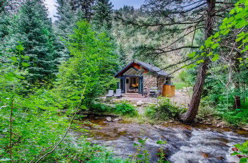 A cozy stone cabin nestled among lush green trees by a flowing stream in a serene natural setting.
