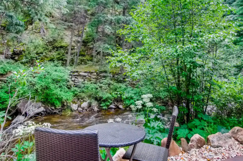 A serene outdoor scene featuring a table and chairs by a flowing stream, surrounded by lush greenery and trees.