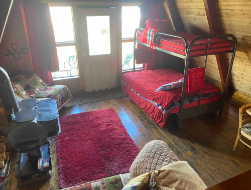 Cozy cabin interior with a bunk bed, red bedding, a wood stove, and a door leading to an outdoor area.