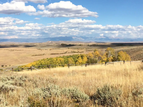 A scenic landscape featuring rolling hills, golden autumn trees, and a blue sky with fluffy clouds.