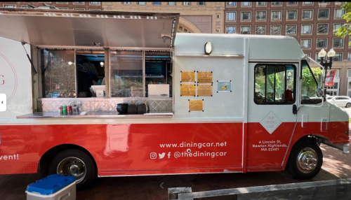 A food truck with a red and white exterior, parked in a city area, featuring a menu and outdoor seating.