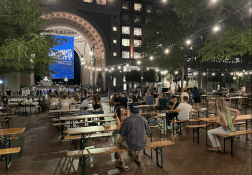 A lively outdoor gathering with people seated at long tables under string lights, enjoying a summer event in the city.