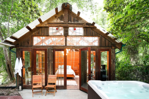 Cozy cabin with large glass doors, wooden accents, and a hot tub, surrounded by lush greenery.