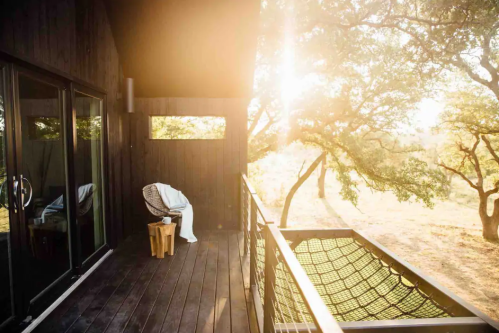 A sunlit balcony with a chair and wooden stool, surrounded by trees and a peaceful landscape.