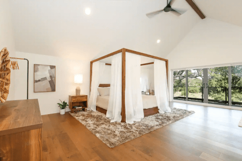 Spacious bedroom with a four-poster bed, soft curtains, wooden furniture, and large windows overlooking greenery.