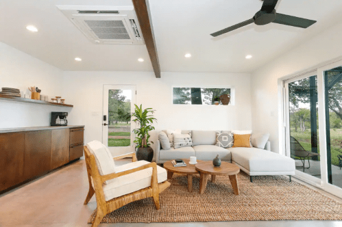 Cozy living room with a gray sofa, wooden coffee table, and large windows overlooking greenery. Modern decor and natural light.