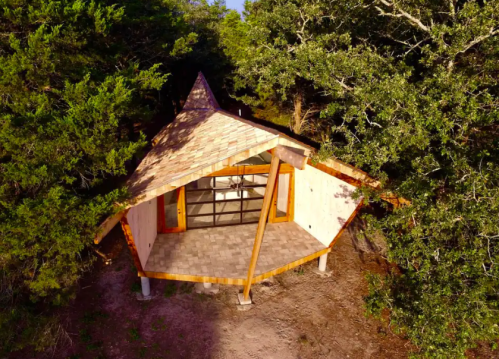 A unique, modern cabin with a pointed roof, surrounded by trees and set on a gravel path.