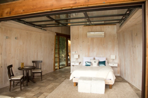 Cozy bedroom with wooden walls, a bed, two nightstands, a small table, and large glass doors opening to the outdoors.