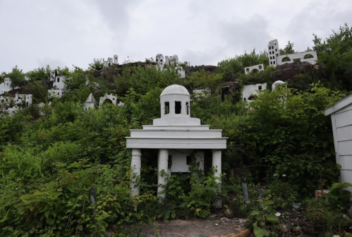 A white structure with columns surrounded by overgrown greenery and small white buildings on a hillside.