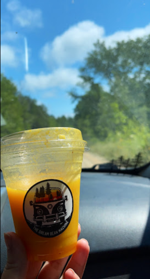 A hand holding a smoothie cup with a logo, set against a sunny, green landscape seen through a car window.