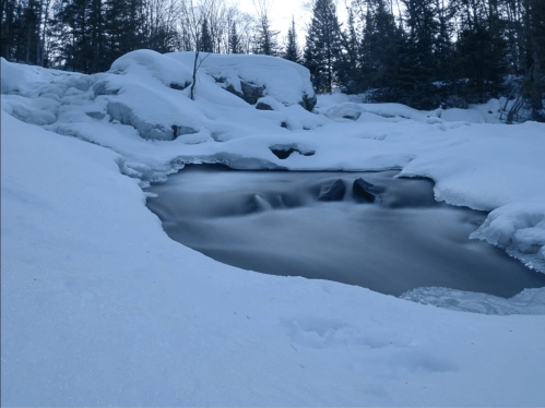 A serene winter scene featuring a partially frozen stream surrounded by snow-covered rocks and trees.