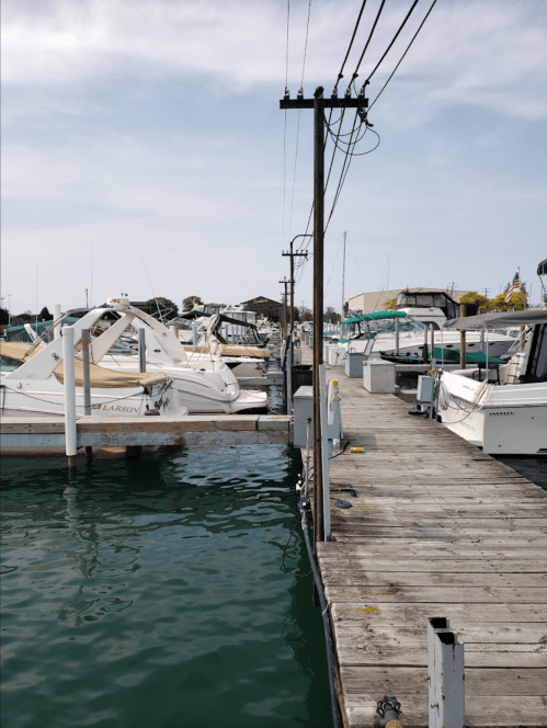 A wooden dock lined with boats, with power lines overhead and calm water reflecting the sky.