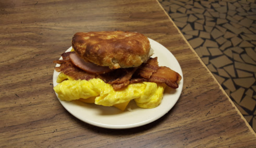 A plate with a biscuit sandwich filled with bacon, ham, and scrambled eggs, served on a wooden table.