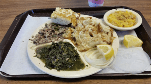 A plate of Southern comfort food featuring greens, black-eyed peas, cornbread, macaroni, and lemon slices.