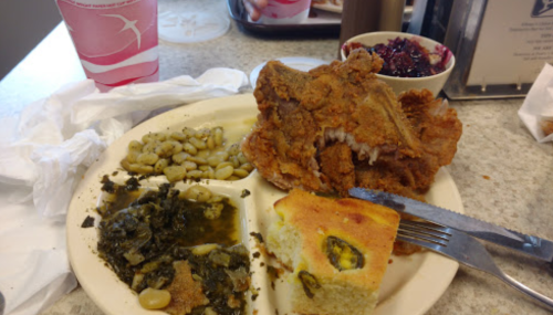 A plate of fried chicken, collard greens, lima beans, cornbread, and a side of berry compote, with a drink in the background.