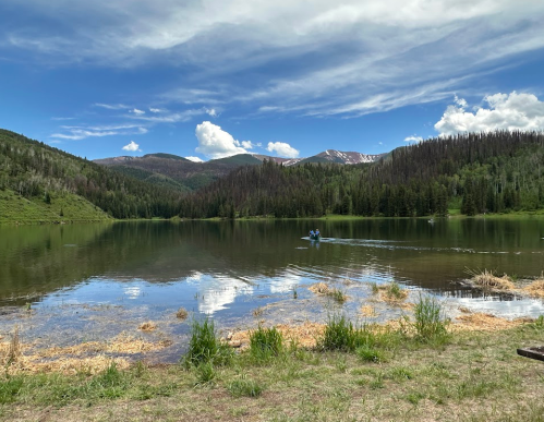 A serene lake surrounded by mountains and trees, with a person kayaking on the water under a blue sky.