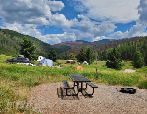 A scenic campsite with tents, a picnic table, and a fire pit, surrounded by green hills and a blue sky with clouds.
